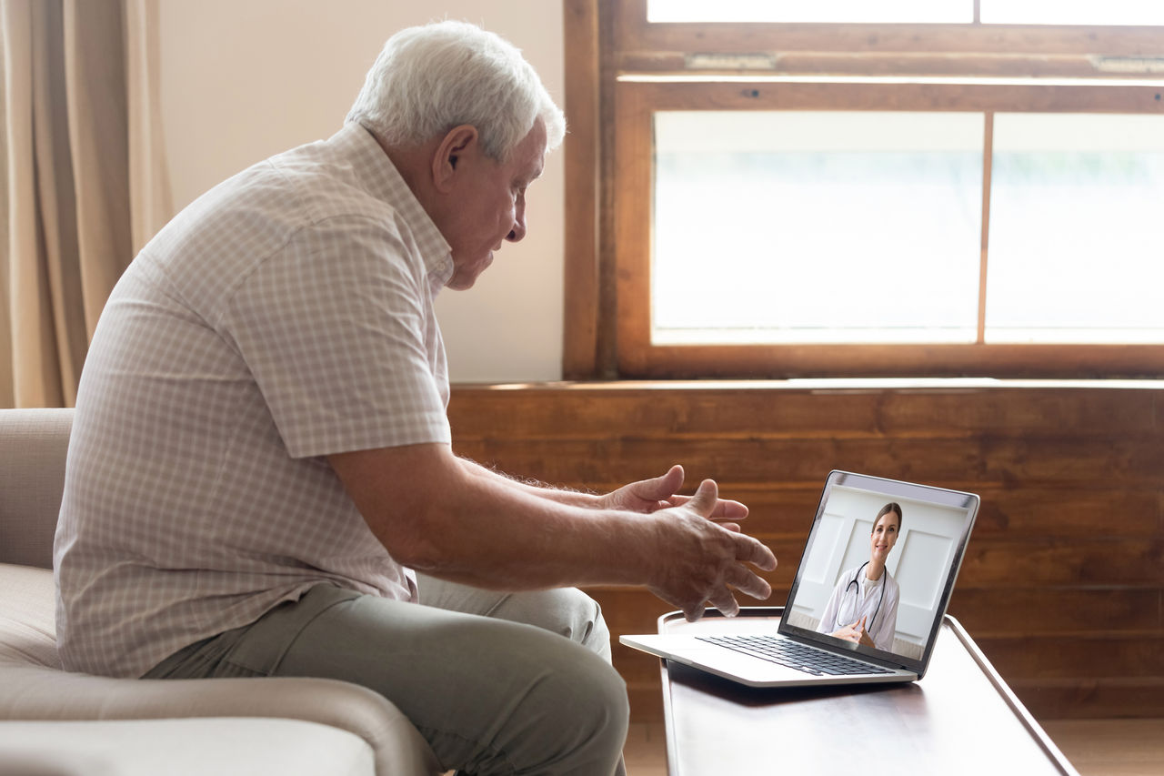 Elderly 70s man seated on sofa make distant video call, senior patient look at laptop screen communicating with doctor therapist online, older generation and modern tech application easy usage concept